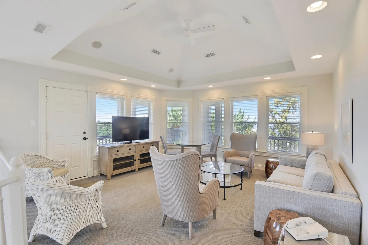 A spacious open-plan living area with a plush sofa, coffee table, and armchairs arranged for conversation or relaxation. Large windows flood the room with natural light, offering glimpses of the ocean and creating a bright, inviting atmosphere. The TV and cozy setup make it perfect for family movie nights, reading, or board games.