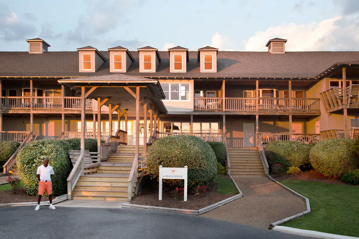 The image features a welcoming porch with bushy landscaping, a sign reading 'Beach House,' and a figure enjoying the outdoor space. The inviting stairs and sunset ambiance suggest a comfortable spot for relaxing after a day of adventure, with plenty of space for pets and owners to unwind together.