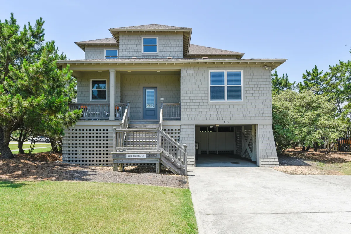 The image showcases the charming exterior of a classic shingle-sided beach house in Duck, North Carolina. The scene centers on a welcoming front porch with inviting steps, surrounded by lush coastal landscaping. The golden light of sunset bathes the porch, hinting at relaxing evenings and ocean breezes. The staircase leads directly to the porch, evoking a sense of arrival and comfort.