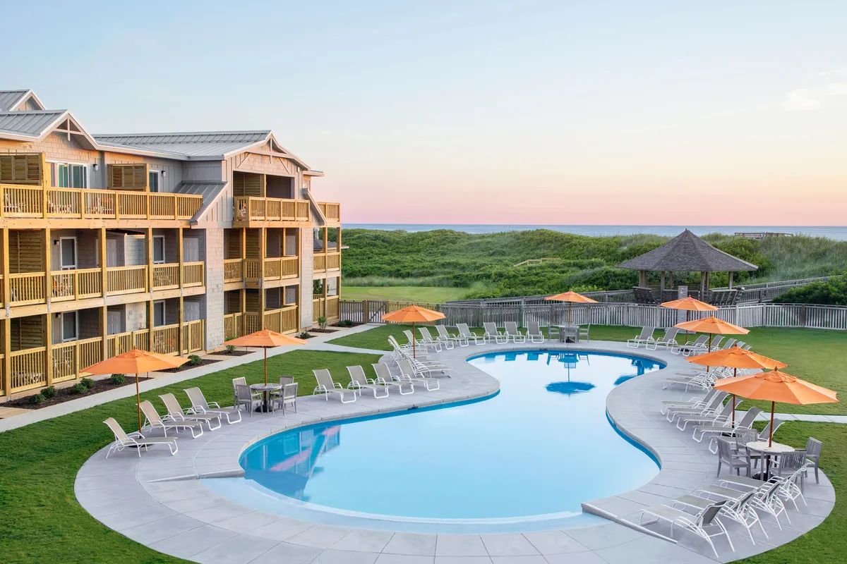 A tranquil outdoor swimming pool framed by a contemporary building facade and an umbrella-shaded seating area. The pool area looks inviting and peaceful, offering both sunny and shaded spots for relaxation. The ambiance suggests an ideal setting for restful swims, sunbathing, or enjoying a quiet morning by the water.