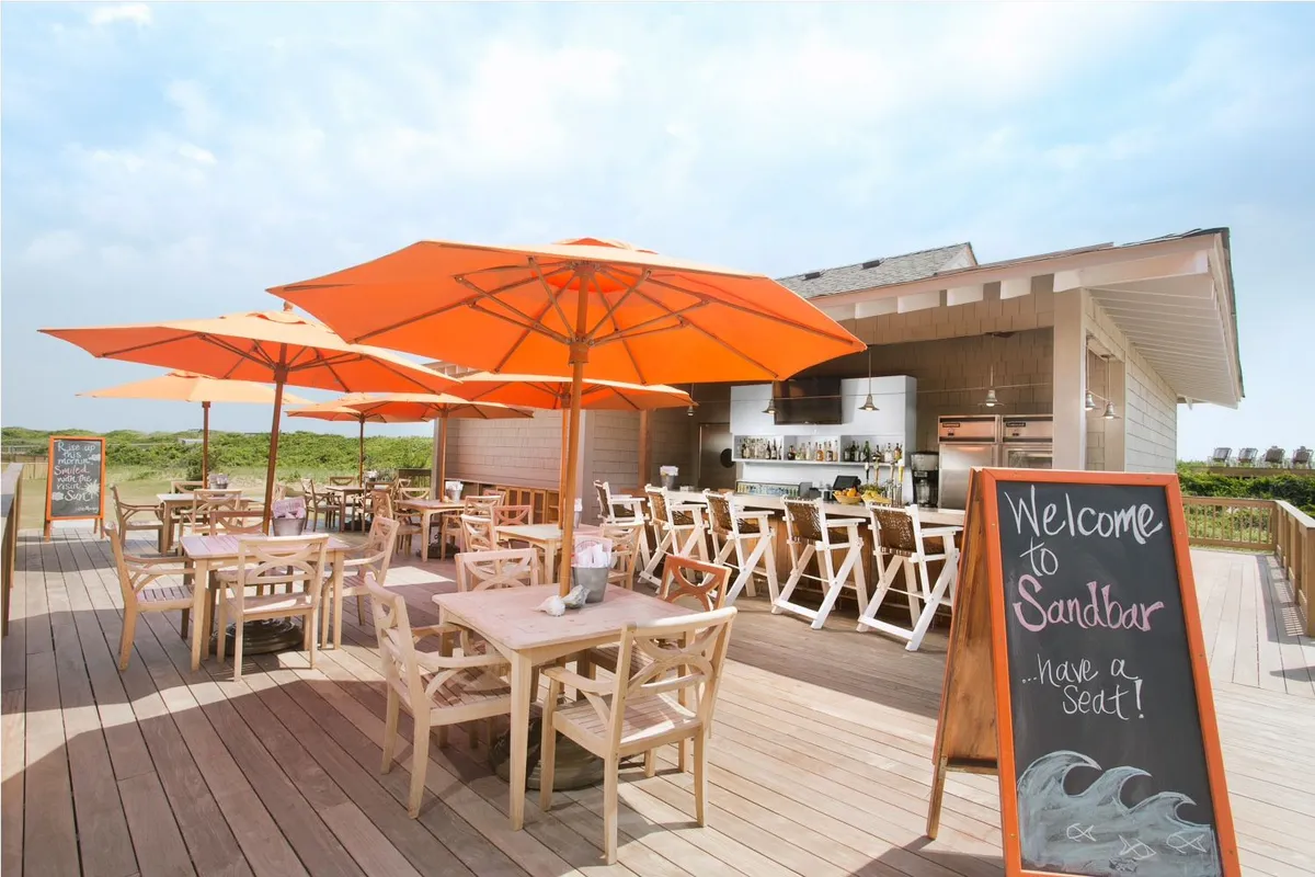 An inviting outdoor dining area with bright orange umbrellas, wooden tables, a bar area with stools, and a visible welcome sign. The ambiance is casual and social, perfect for group meals or snacks after a fun-filled day.