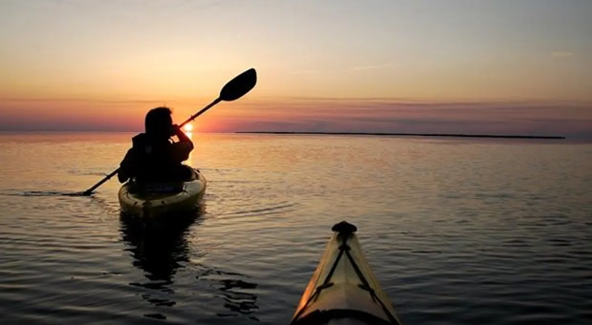 A kayaker glides across calm waters at sunset, with the sky awash in warm colors and the water perfectly mirroring the peaceful scene. The overall mood is serene and contemplative, emphasizing the natural beauty of Currituck Sound at dusk.