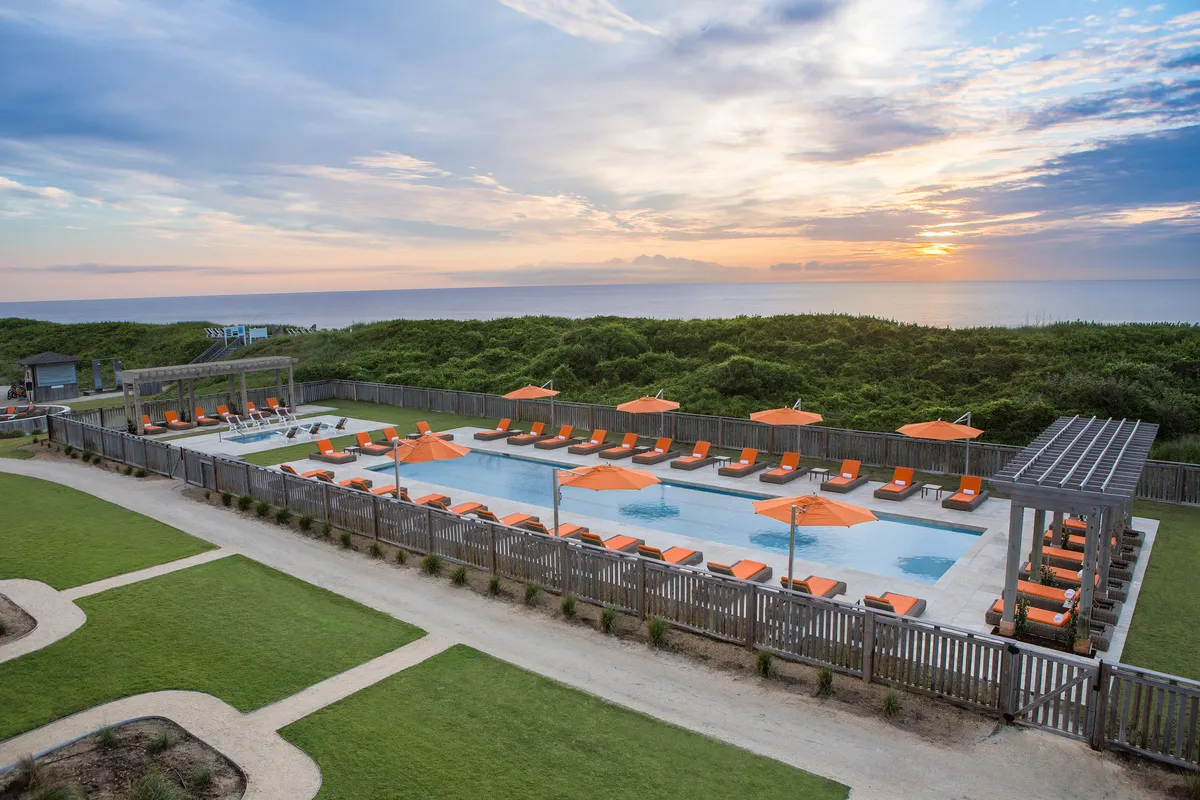 A sparkling swimming pool surrounded by orange umbrellas and lounge chairs, with a beautiful ocean view in the distance. The setting invites both swimming and relaxation, while the tranquil mood hints at social gatherings and evening enjoyment.