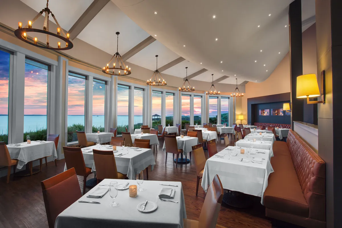 A sophisticated interior dining area with floor-to-ceiling windows offering a spectacular sunset view, elegant table settings, and ambient chandeliers. The serene lighting creates the perfect setting for evening relaxation with a glass of wine.