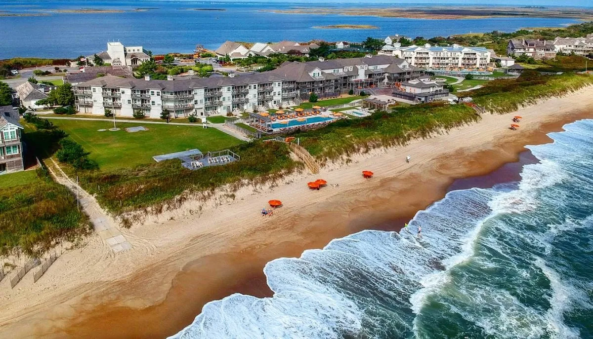 A sweeping view of the beach and ocean waves adjacent to the hotel pool, with vibrant orange umbrellas and a green lawn creating a welcoming transition from poolside to beach. The space is lively yet serene, capturing both pool and beach access in one frame.