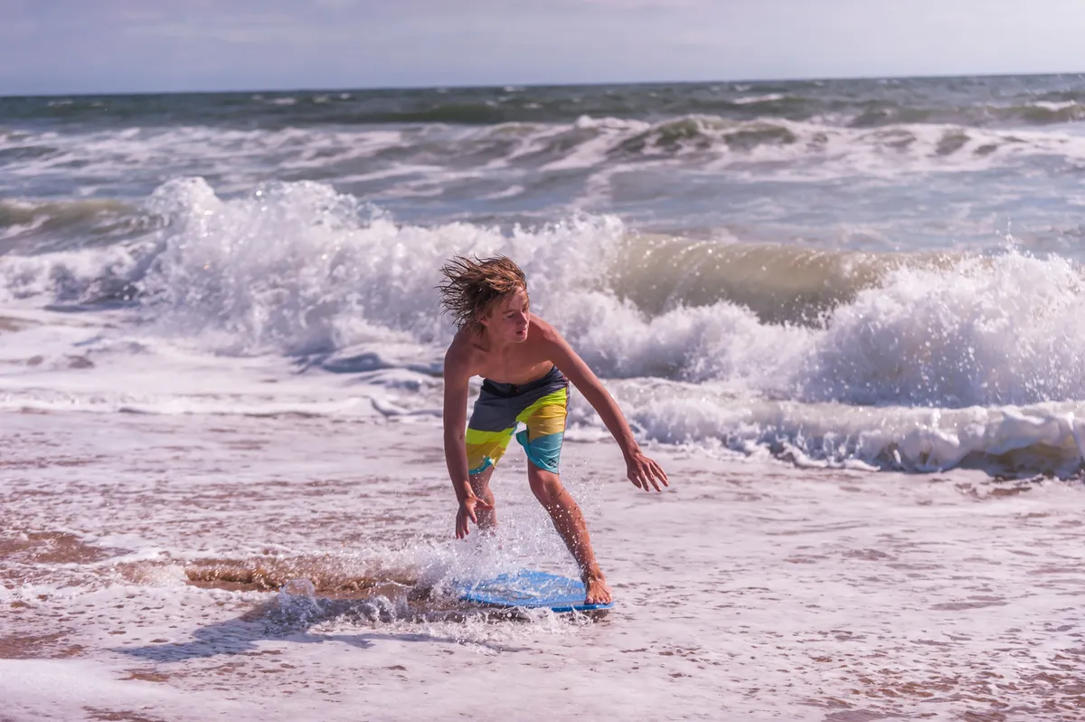 A joyful scene featuring a young boy riding a boogie board on lively ocean waves, with the expansive ocean in the background. The image exudes playful energy and highlights active family fun at the beach.